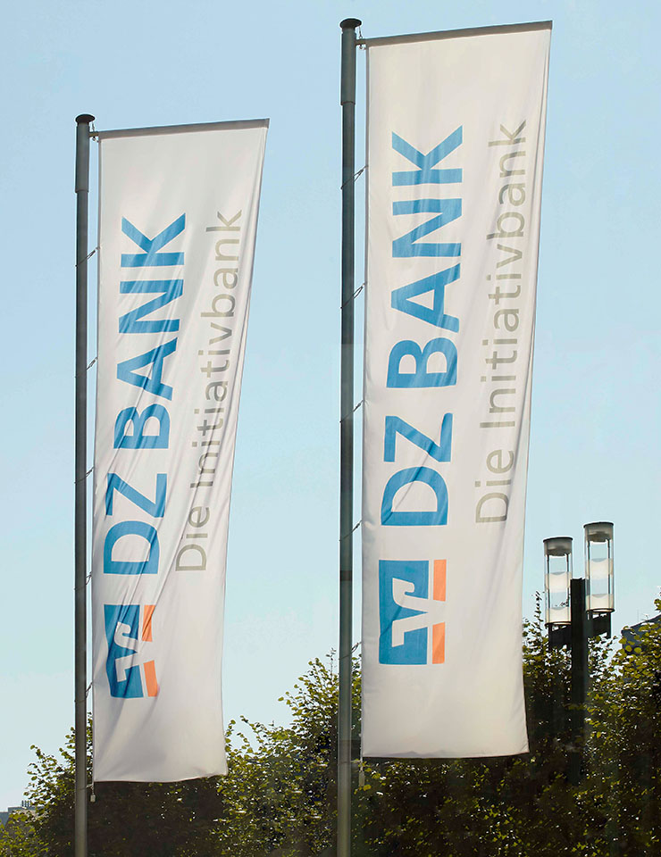 Zwei Fahnen auf denen "DZ Bank - Die Initiativbank" zu lesen ist. Hinter ihnen Himmel, Bäume.