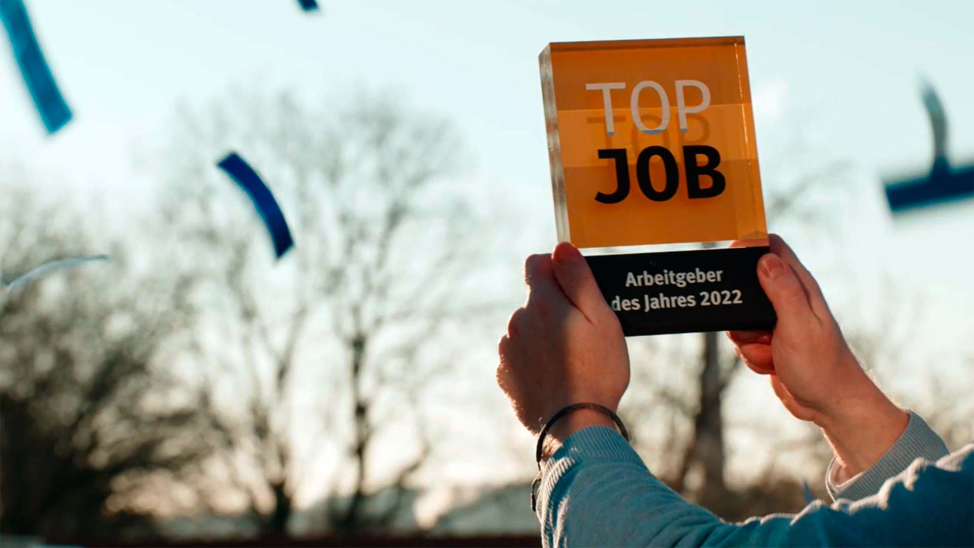 Zwei Hände halten die TOP-JOB-Trophäe in die Höhe. Pokal besteht aus Glas, ist Orange und Schwarz. Auf ihm zu lesen ist: TOP JOB - Arbeitgeber des Jahres 2022. Außen herum fliegt Konfetti.