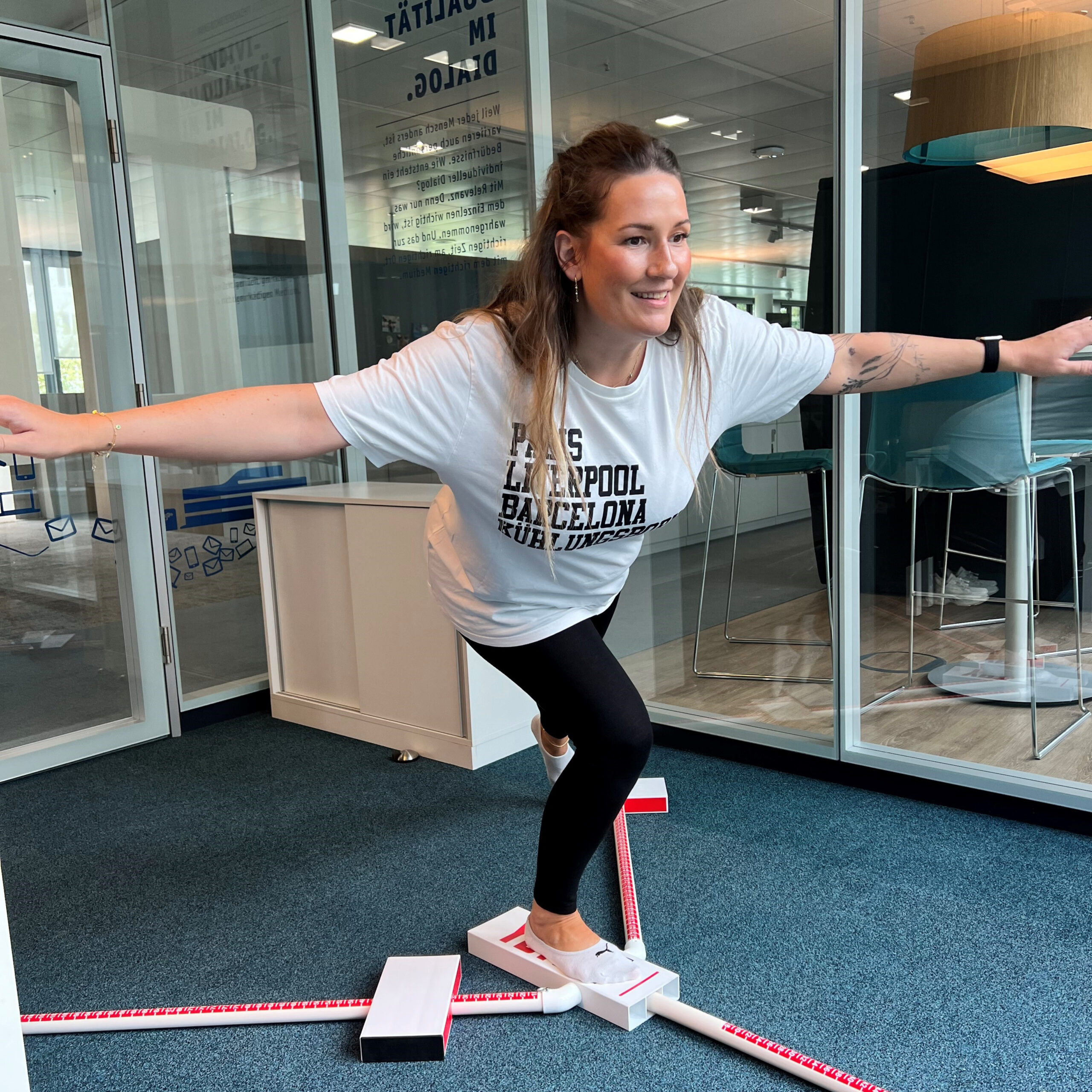 Eine Frau steht in einem Büro balancierend auf einem Bein auf einem weißen Balance-Board mit rot-weißen Markierungen. Sie hat die Arme zur Seite ausgestreckt und lächelt.