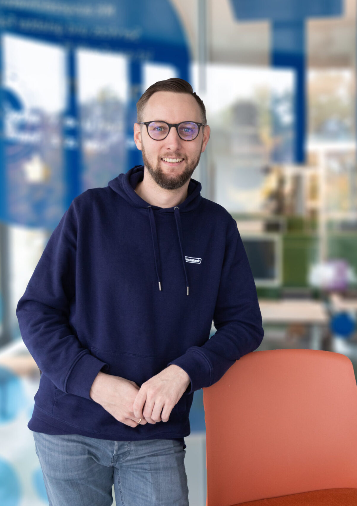 Matthias Zettler steht an einen Stuhl angelehnt im blauen TeamBank-Hoodie und lächelt in die Kamera.
