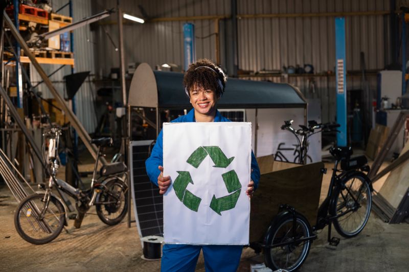 Junge Frau in Blaumann befindet sich in einer Werkstatt und hält ein weißes Papier mit grünem Recyclingzeichen hoch.